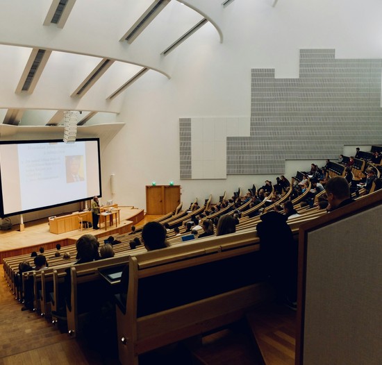 A photo of a university lecture hall looking from the back of a tiered hall down towards the academic speaking.